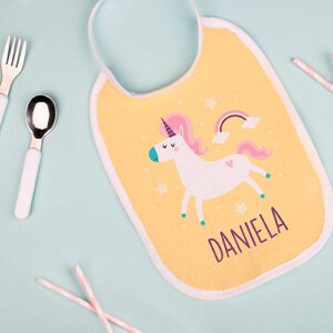 En la imagen, aparece la imagen de un babero personalizado en color amarillo con un unicornio con el nombre de Daniela y junto a un arcoiris. Está apoyado en un entorno azul y junto a él hay unos cubiertos pequeños.